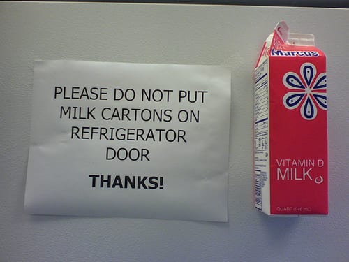 A milk carton is stuck next to a flyer that says, "do not place milk cartons on fridge door."